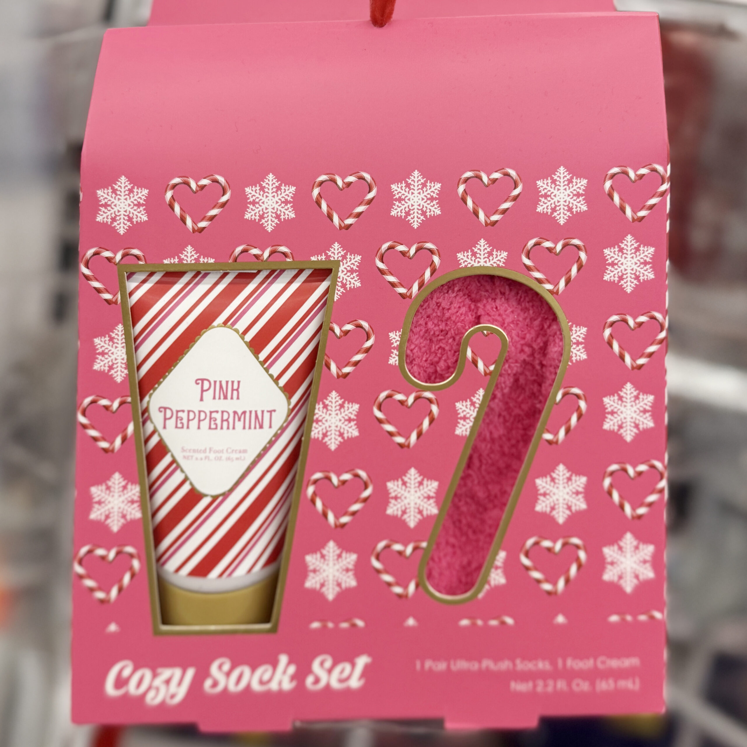 Pink holiday cozy sock set with candy cane and snowflake designs on the packaging. The box displays a striped tube of “Pink Peppermint” foot cream and a candy cane-shaped cutout showing pink plush socks. Text at the bottom reads “Cozy Sock Set.”