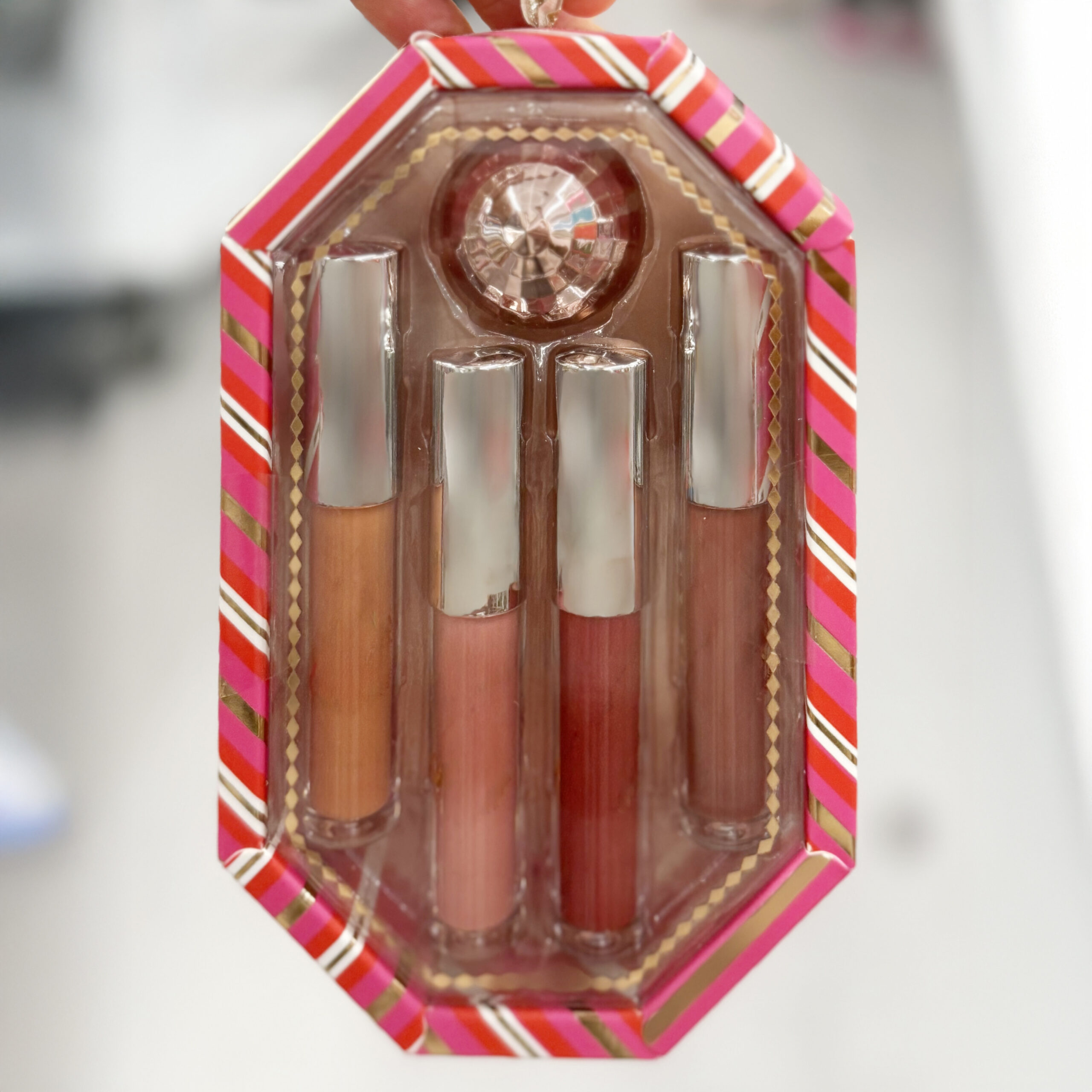 Holiday lip gloss gift set in a pink and red striped octagonal box with a clear packaging window. The set includes four lip gloss tubes in shades of nude, pink, and red, topped with silver caps, and a decorative gem at the top of the packaging.