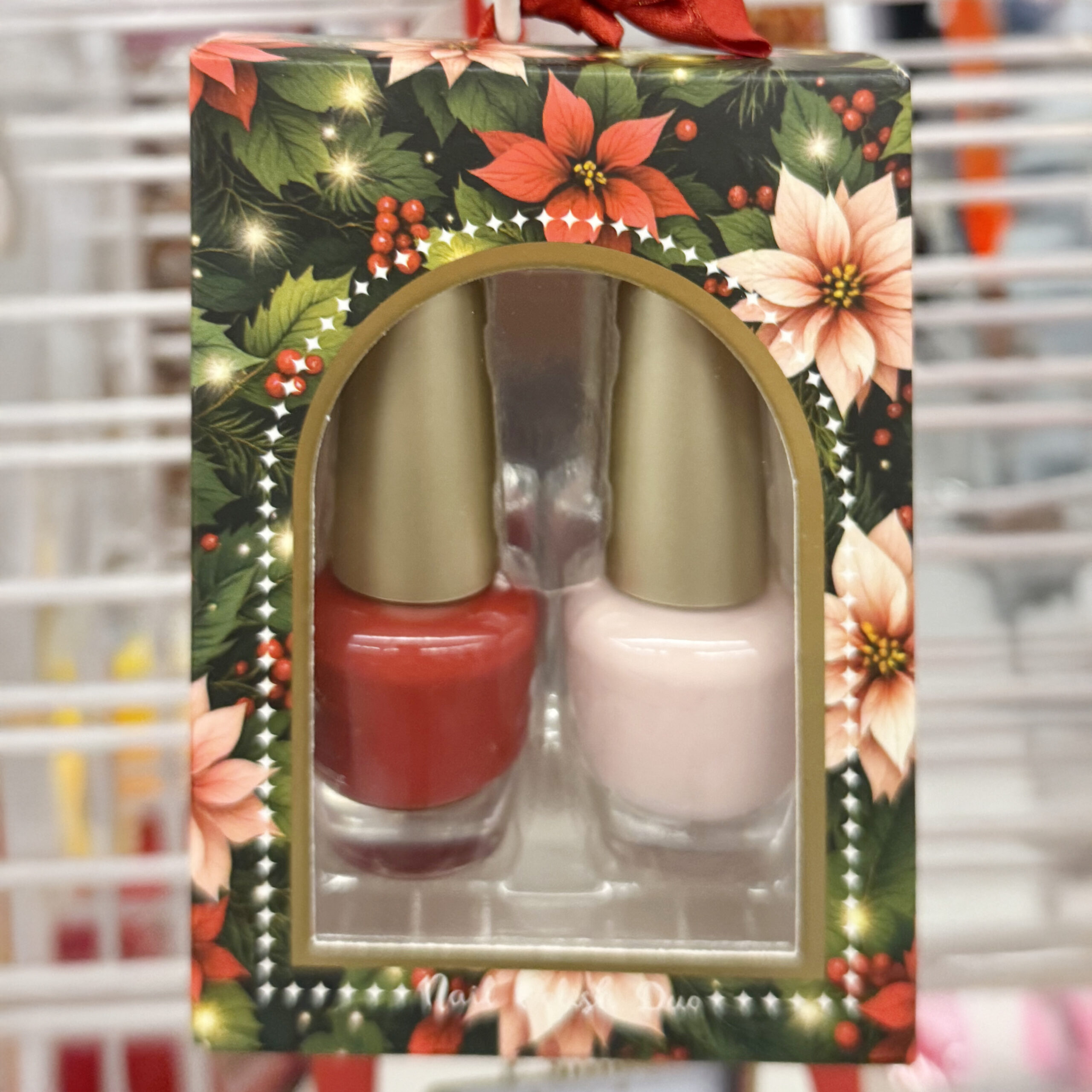 Holiday-themed nail polish duo in a festive floral box decorated with poinsettias and holly. The clear package window reveals two bottles of nail polish with gold caps—one in bright red and the other in pale pink.