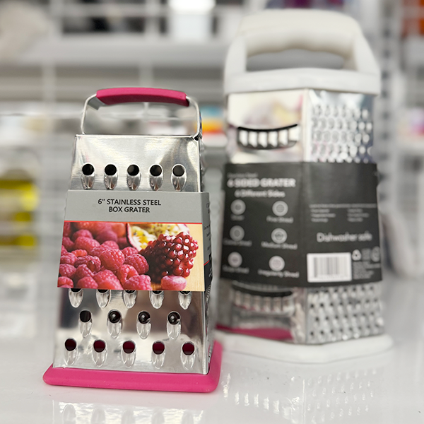 A 6-inch stainless steel box grater with a pink rubber base and handle stands in front of another grater with a white handle on a store shelf.
