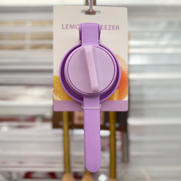A purple manual lemon squeezer is displayed hanging on a card with fruit graphics in the background.