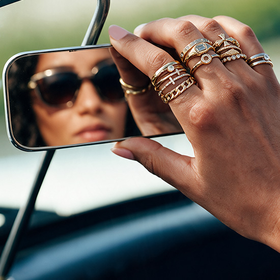 A close-up of a hand wearing multiple stacked gold-tone rings with stone accents while adjusting a rearview mirror.